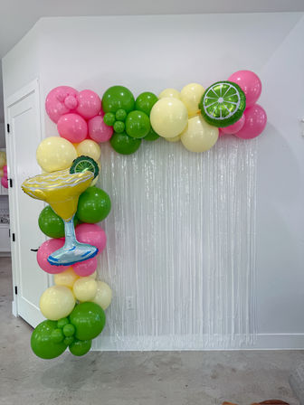 Indoor tropical party balloon arch and garland in pink, green and pale yellow with margarita and lime foil balloons and a white metallic fringe photo backdrop against a white wall