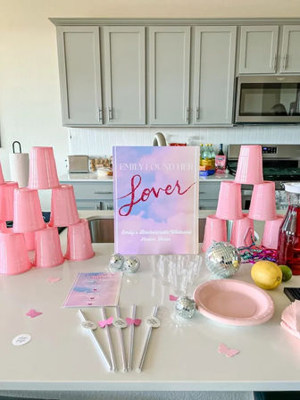 Modern kitchen island styled for a pink bachelorette party with stacked pink plastic cups, pastel “Lover” sign, mini disco balls, clear flutes, pink plate, party straws and a drink pitcher.
