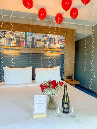 Romantic anniversary hotel room in Austin with red helium balloons floating from the ceiling, white bed, vase of red roses and baby’s breath, champagne bottle and two glasses, and a “Happy Anniversary” card in front of Austin, Texas-themed wall art.
