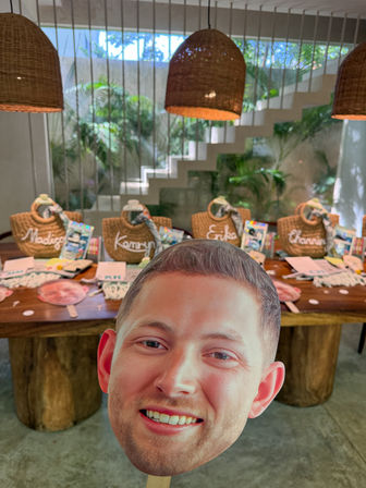 Cheerful oversized face cutout on a stick in the foreground with a wooden welcome table behind, featuring woven straw gift bags, name tags, treats, wicker pendant lights and tropical plants in a bright indoor resort-style space.