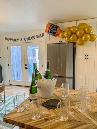 Home kitchen party setup with “WHISKEY & CIGAR BAR” banner over glass doors, gold balloon cluster and inflatable whiskey bottle above fridge, ice bucket with wine bottles and boot-shaped mugs on a wooden island.