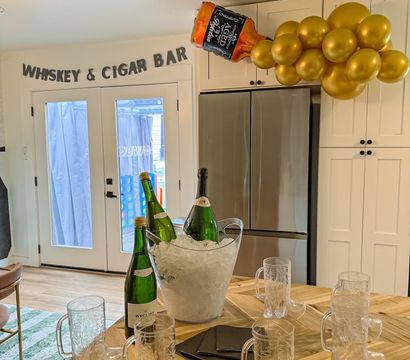 Home kitchen party setup with “WHISKEY & CIGAR BAR” banner over glass doors, gold balloon cluster and inflatable whiskey bottle above fridge, ice bucket with wine bottles and boot-shaped mugs on a wooden island.