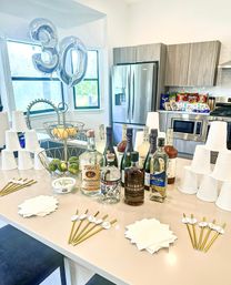 Bright modern kitchen set up as a 30th birthday cocktail bar with silver "30" balloons, assorted liquor bottles and mixers on a countertop, stacked white cups, napkins, gold stirrers, fruit bowls and stainless steel refrigerator in the background.