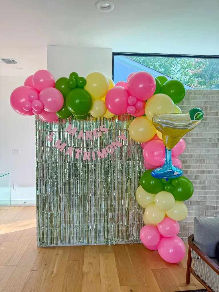 Indoor party photo backdrop with metallic fringe curtain, pink, green and yellow balloon garland and a large margarita-shaped foil balloon; pink letters spell “Margs & Matrimony”.