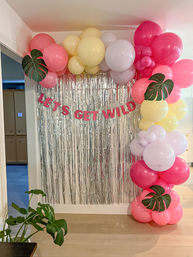 Indoor party photo backdrop with silver fringe curtain, pink banner reading "LET'S GET WILD", and a colorful balloon arch in pink, yellow, and lavender accented with faux monstera leaves.