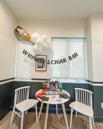 Cozy indoor whiskey and cigar bar corner with a banner, white balloons and inflatable beer bottle above a small round table stocked with whiskey bottles, mixers, napkins and playing cards between two white chairs.