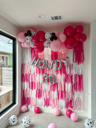 Cheerful pink birthday backdrop: layered pink-and-white fringe wall topped with hot pink, light pink and black‑spotted balloons and silver foil "HOWDY 30" balloons, indoor home party decor