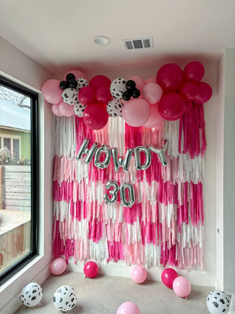 Cheerful pink birthday backdrop: layered pink-and-white fringe wall topped with hot pink, light pink and black‑spotted balloons and silver foil "HOWDY 30" balloons, indoor home party decor