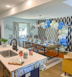 Bright open-plan kitchen and dining room styled for a party with silver "MATCH MADE IN HEAVEN" letter balloons, blue-and-white balloon arch, long table set with holographic gift bags, blue caps and playful face cutouts, and a decorated kitchen island with snacks and photos.