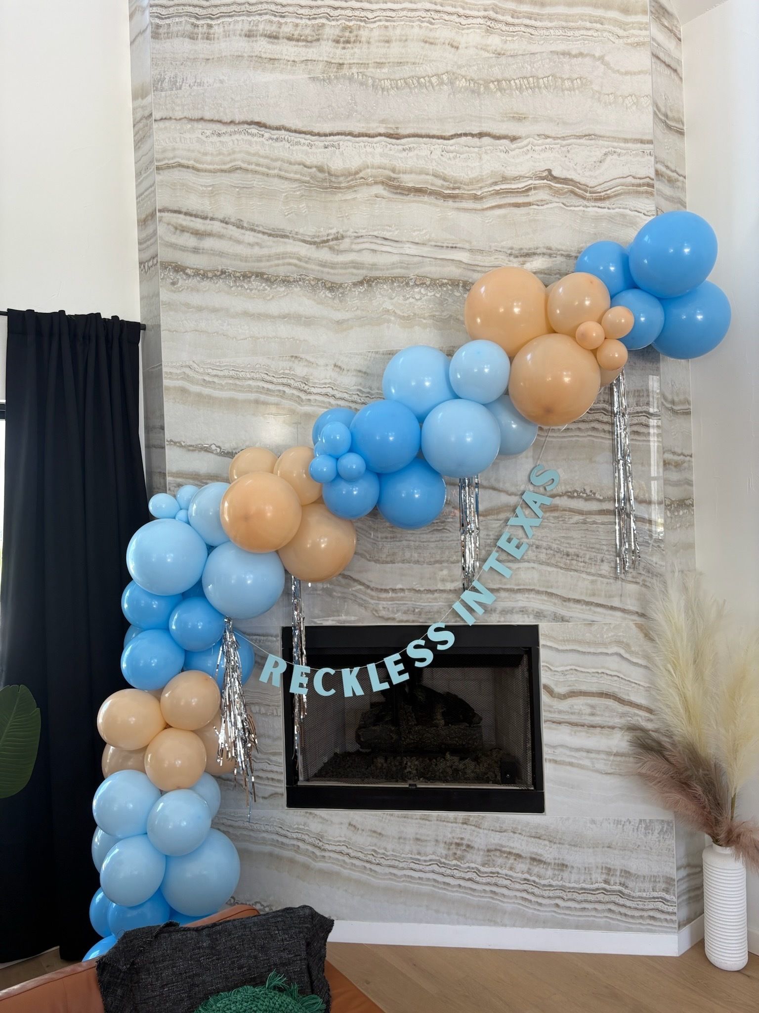 Playful blue and tan balloon garland with silver tassels draped over a modern marble fireplace, featuring a "RECKLESS IN TEXAS" banner, pampas grass and dark curtains — living room party decor in Texas.