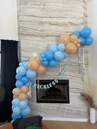 Playful blue and tan balloon garland with silver tassels draped over a modern marble fireplace, featuring a "RECKLESS IN TEXAS" banner, pampas grass and dark curtains — living room party decor in Texas.