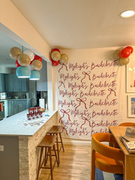 Open-plan modern kitchen decorated for a bachelorette party with a custom script backdrop and bow motifs, clusters of red and gold balloons, teal pendant lights, countertop set with red cups and confetti, wooden bar stools and orange dining chairs.