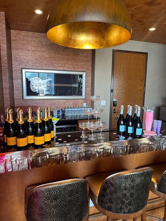 Hotel-suite bar setup for a celebration: wood counter with multiple champagne bottles, stacked coupe glass tower, bottled water, pink cups and metallic fringe under a large brass pendant lamp, two patterned bar stools.