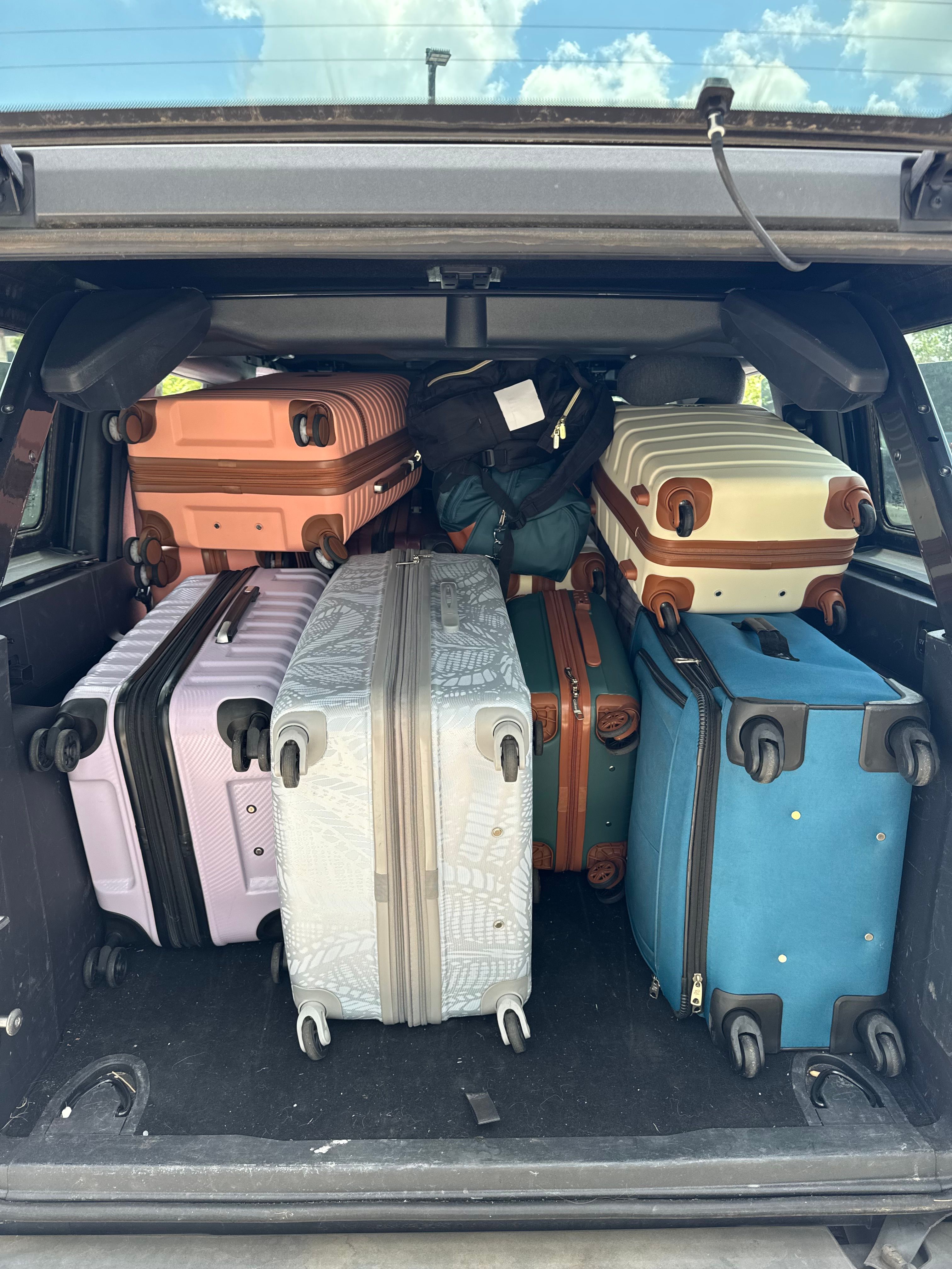 Open SUV trunk filled with stacked colorful wheeled suitcases and a backpack, packed and ready for a road trip or airport travel under a blue sky