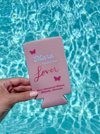Cheerful poolside shot of a manicured hand holding a pink personalized bachelorette can cooler above sparkling turquoise pool water.