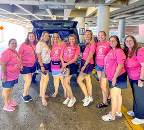 Smiling group of women in matching pink shirts posing at an airport passenger pick-up curb beside an open SUV trunk with luggage.
