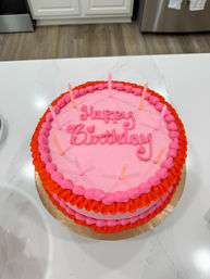 Bright pink round birthday cake with red and hot-pink piped ruffle border and "Happy Birthday" script, topped with slim pink candles, sitting on a white kitchen island countertop