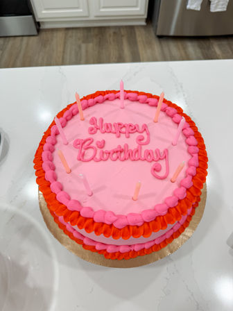 Bright pink round birthday cake with red and hot-pink piped ruffle border and "Happy Birthday" script, topped with slim pink candles, sitting on a white kitchen island countertop