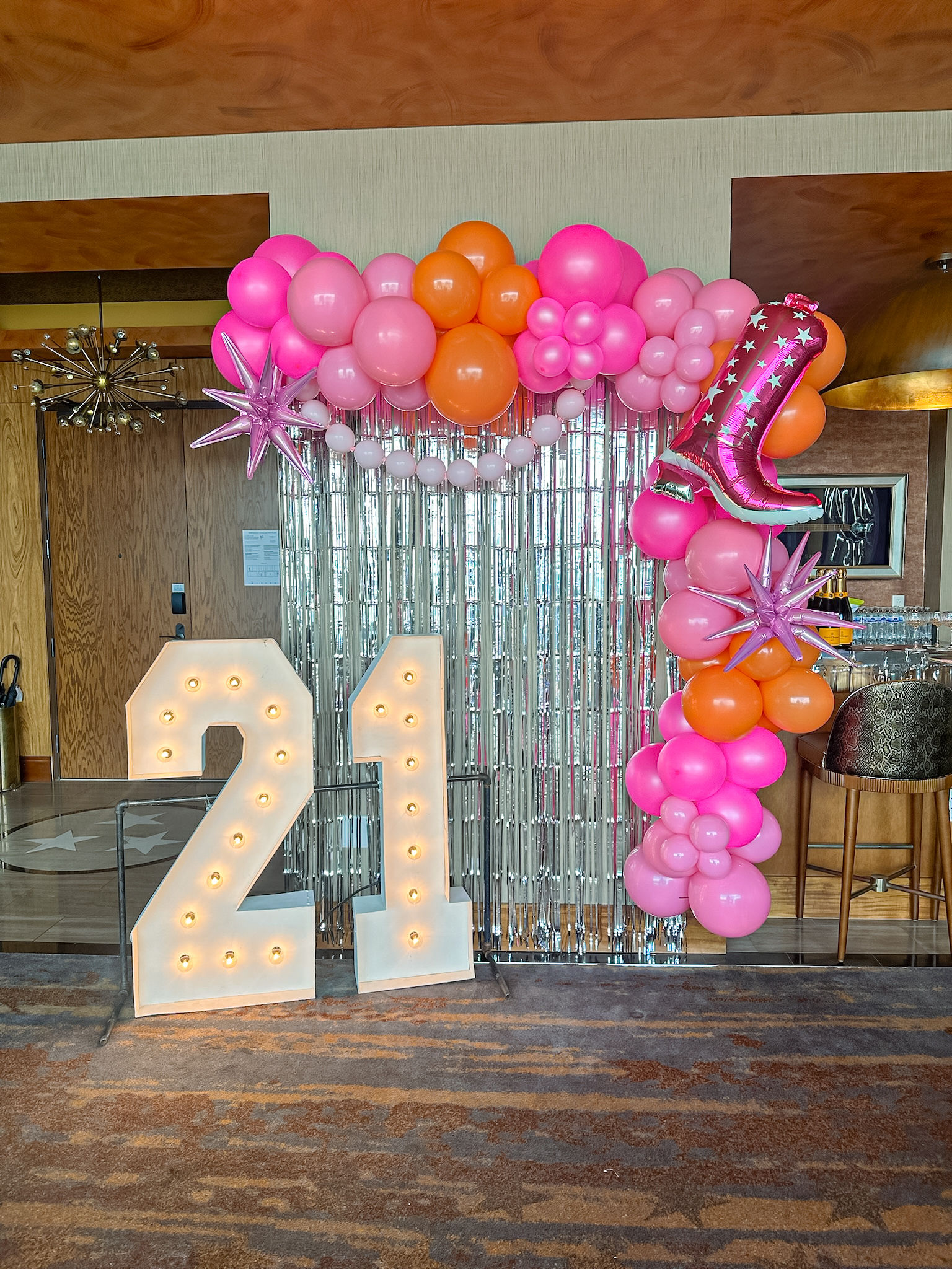 Indoor event photo backdrop for a 21st birthday: illuminated white marquee "21" numbers, pink and orange balloon garland with starburst and cowboy-boot foil balloons, silver fringe curtain