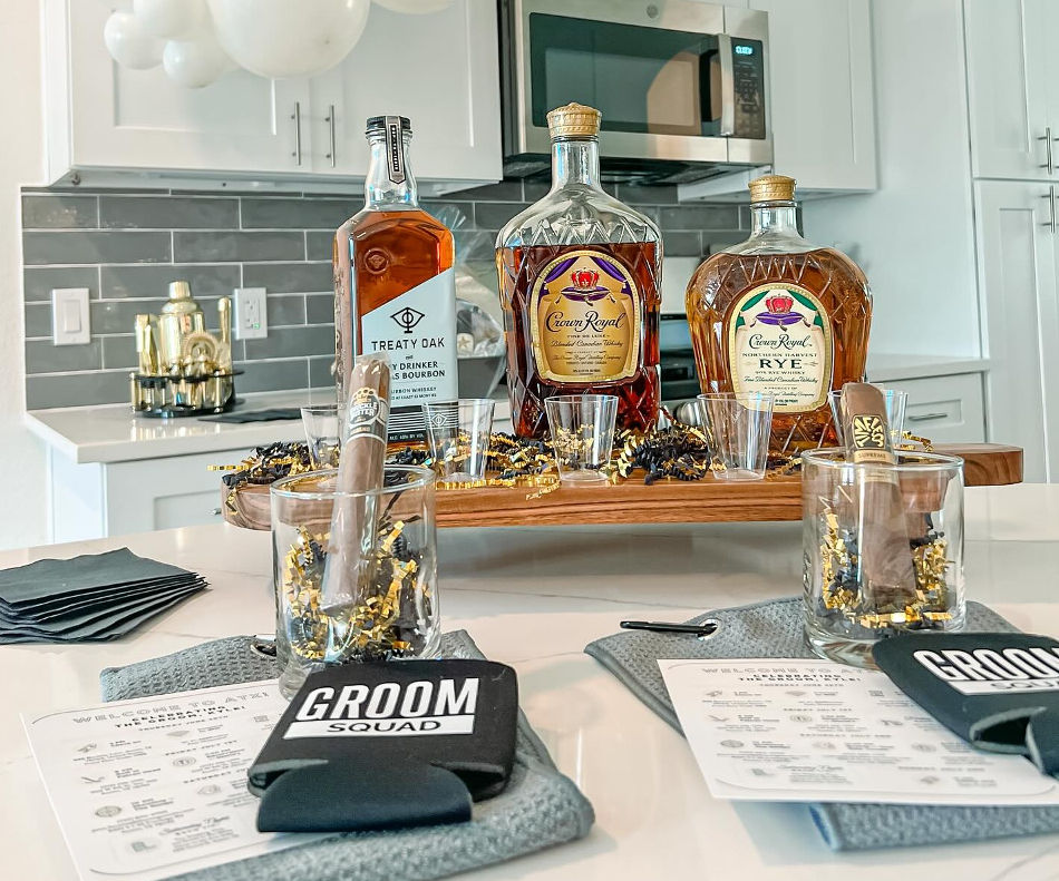 Groom Squad bachelor-party spread on a modern kitchen island: three whiskey bottles, cigars in rocks glasses, shot glasses, black koozies, and gold confetti party decor.