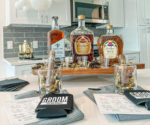 Groom Squad bachelor-party spread on a modern kitchen island: three whiskey bottles, cigars in rocks glasses, shot glasses, black koozies, and gold confetti party decor.