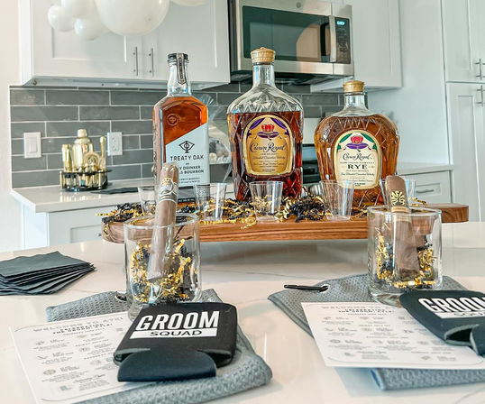 Groom Squad bachelor-party spread on a modern kitchen island: three whiskey bottles, cigars in rocks glasses, shot glasses, black koozies, and gold confetti party decor.