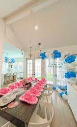 Open-plan dining room party setup with a long wooden table lined with bright pink fluffy cowboy hats, white chairs, blue-and-white balloon arch and shimmering fringe backdrop by French doors to a backyard pool.