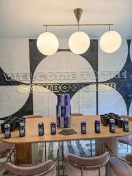 Contemporary indoor lounge with geometric wall mural and neon sign, three globe pendant lights above a wooden table staged for a party with stacked beer cans, black cups, napkins, and pink velvet chairs.