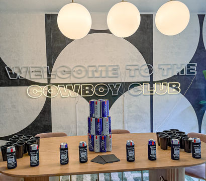Contemporary indoor lounge with geometric wall mural and neon sign, three globe pendant lights above a wooden table staged for a party with stacked beer cans, black cups, napkins, and pink velvet chairs.