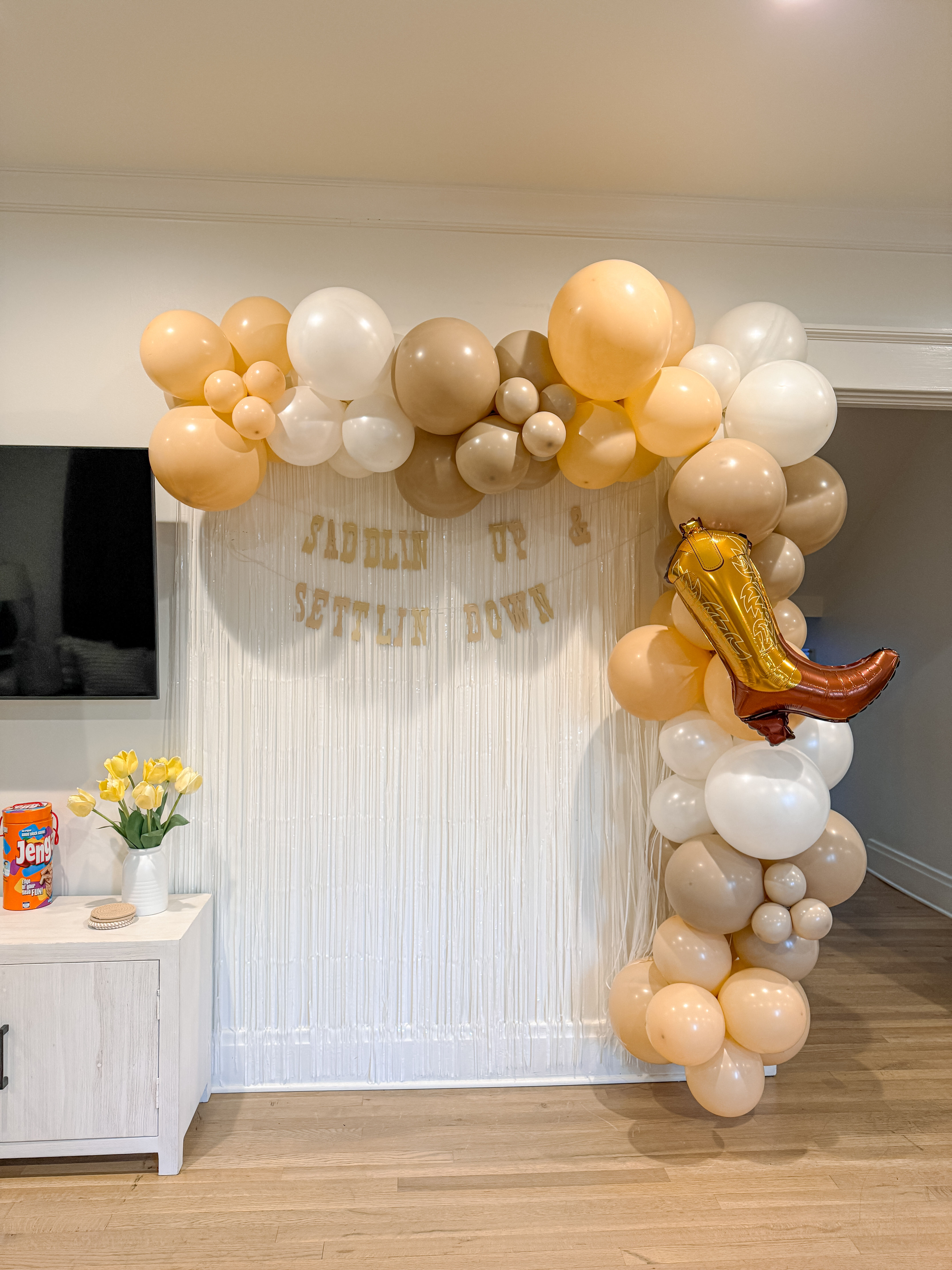Indoor living-room party photo backdrop with beige, cream, and white balloon arch and bronze cowboy-boot foil balloon draped over a white fringe curtain; small side table with yellow tulips and boxed game.