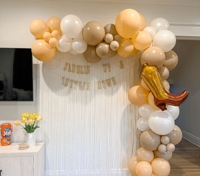 Indoor living-room party photo backdrop with beige, cream, and white balloon arch and bronze cowboy-boot foil balloon draped over a white fringe curtain; small side table with yellow tulips and boxed game.
