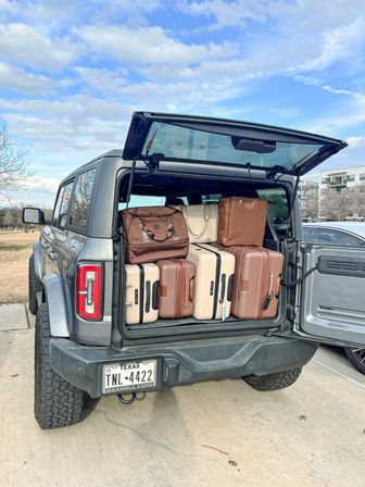 Gray SUV with open tailgate revealing neatly packed brown and beige suitcases and travel bags, ready for a road trip in a suburban Texas parking lot under a bright blue sky.