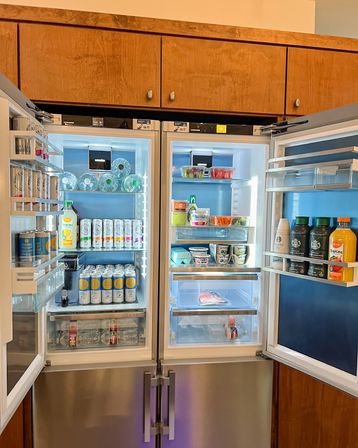 Open modern stainless-steel French-door refrigerator in a kitchen with wooden cabinets, brightly lit and neatly stocked with bottled water, canned drinks, juice, yogurt tubs, eggs, pre-cut produce and condiments.