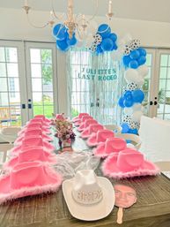 Fun bachelorette party scene: long rustic table lined with pink fluffy cowboy hats, a white rhinestone “BRIDE” cowboy hat at the head, floral centerpiece, and a blue-and-white balloon arch with silver fringe backdrop in a bright indoor dining room.