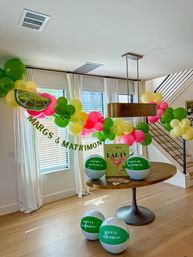 Cheerful margarita-themed indoor party setup with lime-slice and green, pink, yellow balloon garland over a modern round wood table, matching beach balls and party sign in a sunlit dining area with white curtains and staircase.