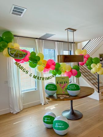 Bright tropical-themed indoor party in a modern dining room — lime-slice and green, yellow, pink balloon garland with matching beach balls and a 'MARGS & MATRIMONY' banner over a round wood table beneath a pendant light.