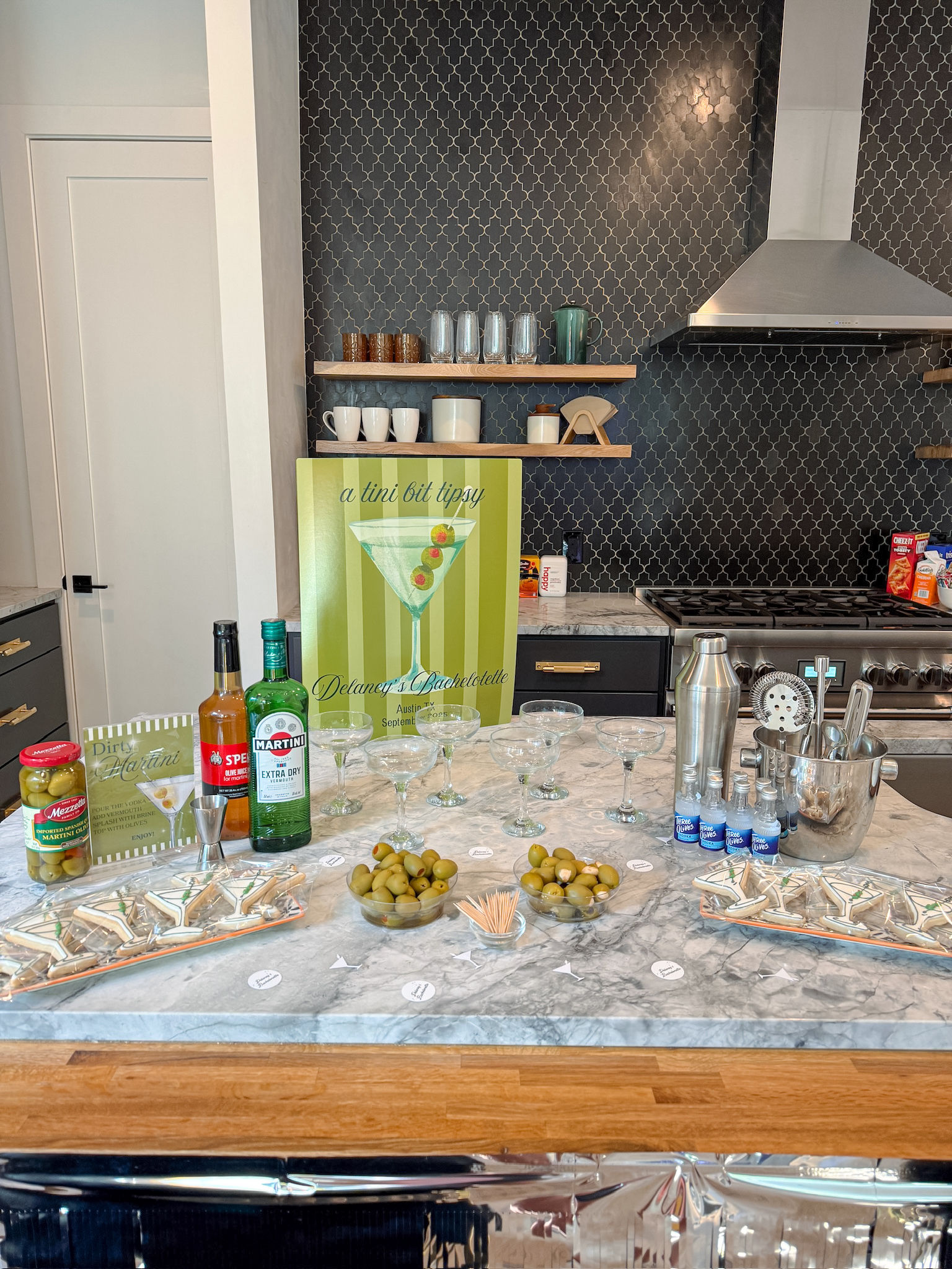 Modern kitchen island cocktail station with a green martini sign, rows of coupe glasses, bottles and a metal shaker, bowls of green olives and martini-shaped cookies on a marble countertop against a dark patterned backsplash.