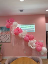 Cheerful pink and white balloon garland with bow accents and long streamers draped across a pink interior wall above a round dining table and window