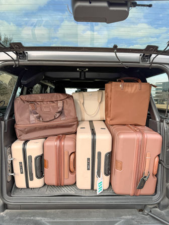 Open SUV trunk packed with beige and terracotta hard-shell suitcases, a brown duffel and tote neatly stacked—ready for a road trip with an urban parking backdrop.