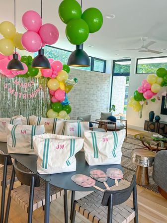Bright modern living room styled for an indoor celebration with pink, green and yellow balloon garlands, metallic fringe backdrop, black pendant lights, a table of monogrammed canvas tote gift bags and face-cutout photo props.