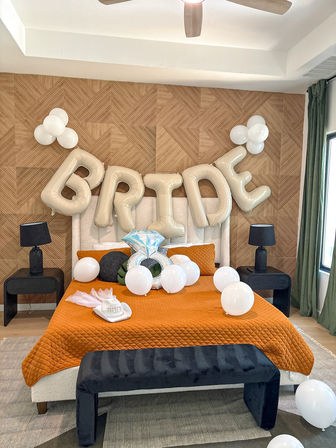 Modern bedroom staged for a bridal celebration with large 'BRIDE' gold balloons above an orange quilted bedspread, white balloons, wood accent wall and black lamps