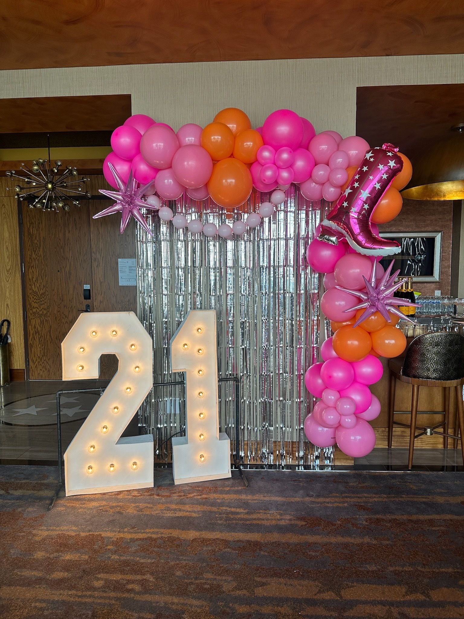 Bright 21st birthday backdrop: lit marquee '21' numbers, pink and orange balloon arch with star and boot foil balloons, silver fringe curtain in an indoor event space.