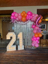 Bright 21st birthday backdrop: lit marquee '21' numbers, pink and orange balloon arch with star and boot foil balloons, silver fringe curtain in an indoor event space.