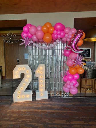 Bright 21st birthday backdrop: lit marquee '21' numbers, pink and orange balloon arch with star and boot foil balloons, silver fringe curtain in an indoor event space.
