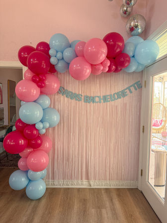 Pink indoor bachelorette backdrop with pink and blue balloon garland, metallic fringe curtain, light-blue banner reading “Bachelorette,” disco ball accents and wood floor