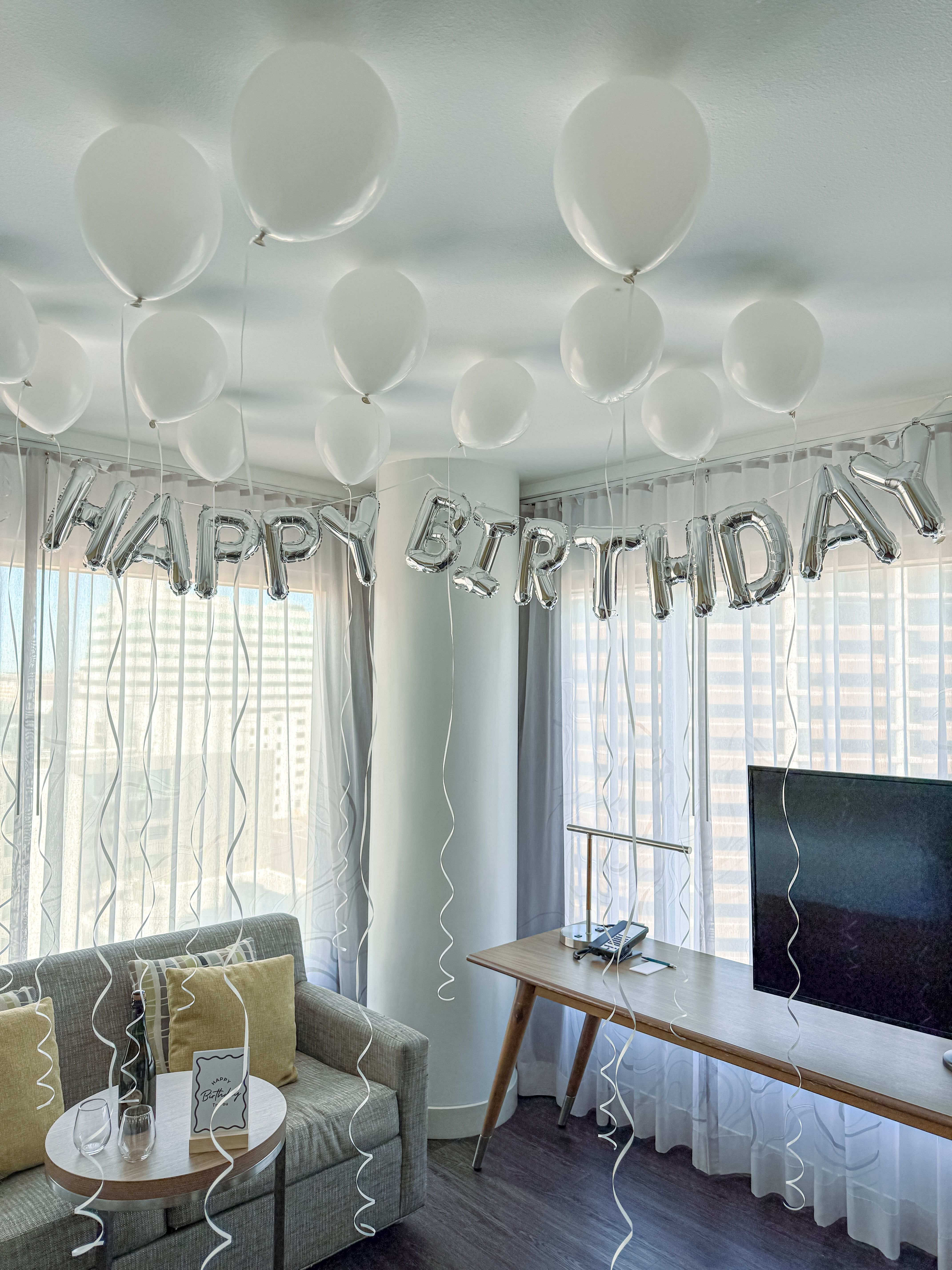 White helium balloons floating to the ceiling and silver "HAPPY BIRTHDAY" foil letters strung across a bright hotel-suite living area with sofa, side table, desk and city-view floor-to-ceiling windows.