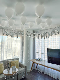 White helium balloons floating to the ceiling and silver "HAPPY BIRTHDAY" foil letters strung across a bright hotel-suite living area with sofa, side table, desk and city-view floor-to-ceiling windows.