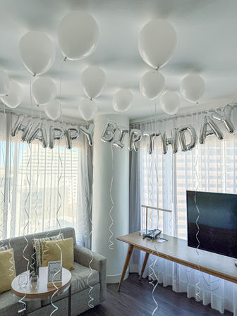 White helium balloons floating to the ceiling and silver "HAPPY BIRTHDAY" foil letters strung across a bright hotel-suite living area with sofa, side table, desk and city-view floor-to-ceiling windows.