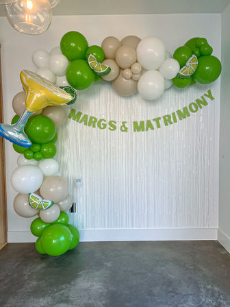 Margarita-themed party backdrop with green, white and beige balloon garland, lime-slice accents and a foil margarita glass balloon framing a 'MARGS & MATRIMONY' banner over a white fringe backdrop.