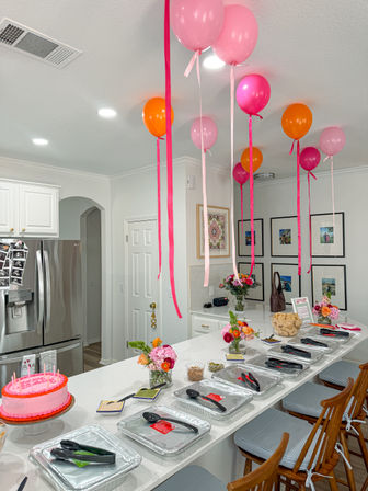 Bright modern home kitchen party setup with pink and orange balloons and ribbons over a white island lined with trays, a pink cake, flower arrangements and bar stools.